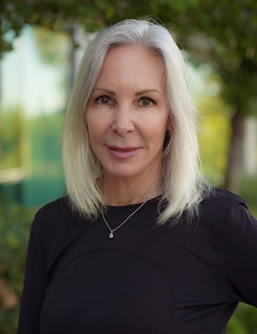 Portrait of Debra Gromacki, Aesthetic Injector, with straight shoulder-length silver hair, wearing a black top and necklace, standing outdoors with greenery in the background