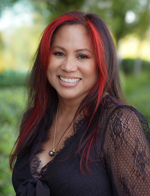 Portrait of Ilsa Ramos, MA, smiling outdoors with long dark hair and red highlights
