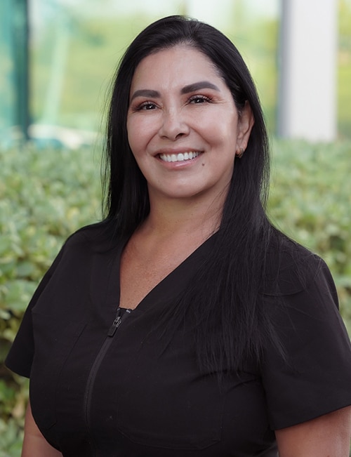 Lupe, a Nurse Practitioner specializing in aesthetic medicine, smiling outdoors in front of greenery, wearing a black zip-up top