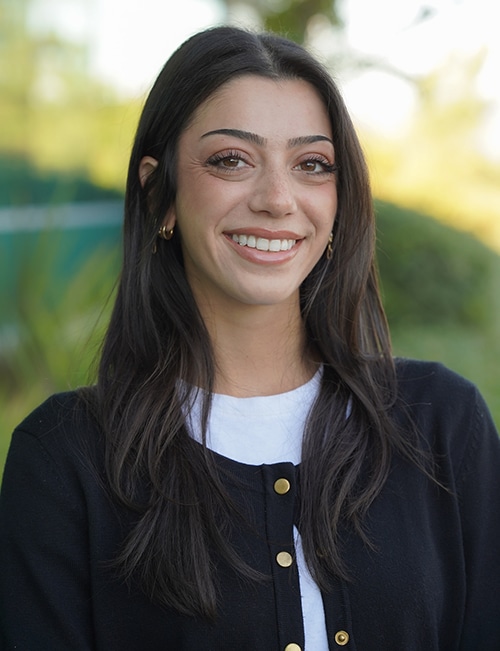 Danielle Hanna, Social Media Manager, smiling outdoors with long dark brown hair, wearing a black sweater with gold buttons