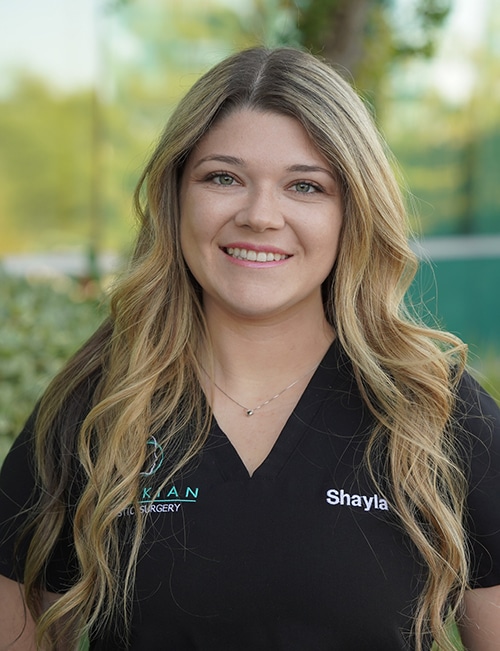 Shayla Jackson, MA, Medical Assistant, smiling outdoors with long wavy blonde hair, wearing a black scrub top embroidered with her name