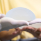 A plastic surgeon wearing white gloves holds a silicone breast implant during a consultation, with a patient in a yellow shirt observing in the background