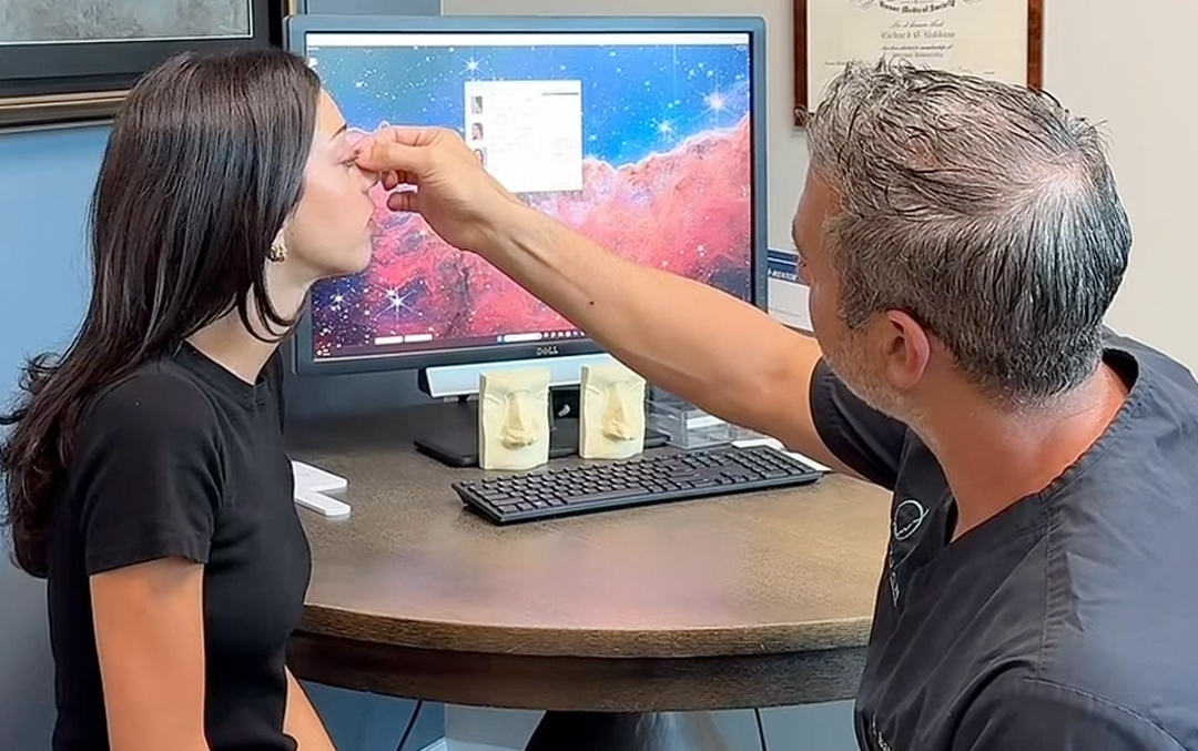 Dr. Richard Balikian examining a patient’s nose during a rhinoplasty consultation, with a computer in the background displaying imaging software.